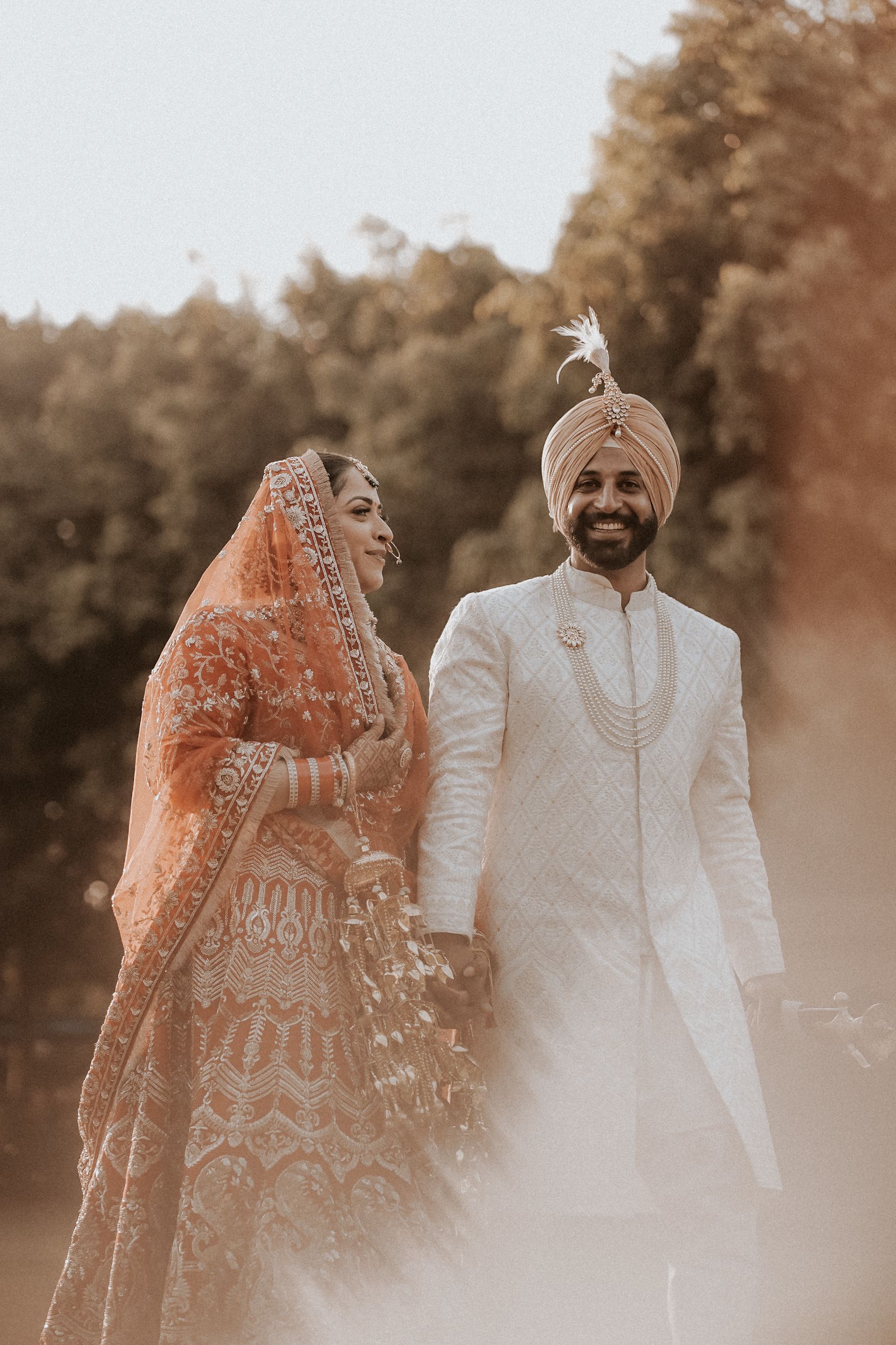 A bride in an embroidered orange veil and a smiling groom in ivory wedding attire, standing together outdoors.